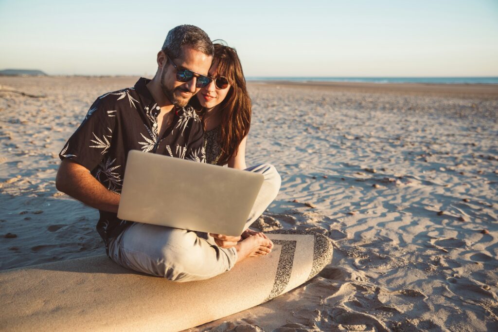 Couple with surfboard sitting on the beach, using laptop